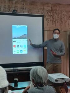 スマホ教室で、投影されたスマホのアプリ画面を指さしながら説明する講師。プロジェクターにはスマホのホーム画面とアプリアイコンが表示されている。