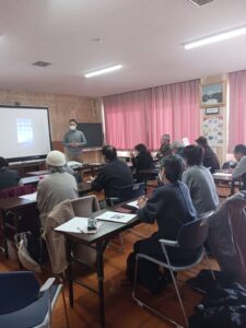 グレーのセーターを着た早川スタイルの講師が、プロジェクターでスマホ画面を投影しながらシニア向けスマホ操作を説明している様子。参加者はピンクのカーテンのある和室風の教室で熱心に聞いている。